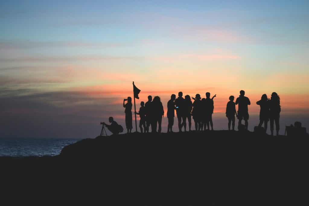 A group of people are shown in silhouette at the top of a mountain with a flag and sunset in background